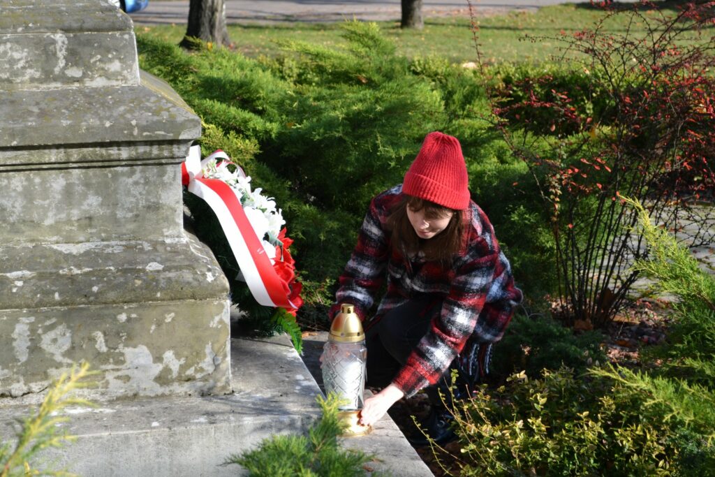 Kobieta w czerwonej czapce kuca. Składa znicz przed pomnikiem.