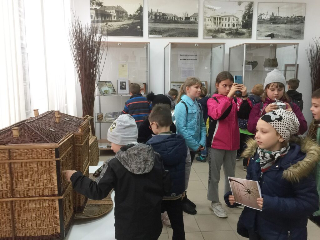 Dzieci oglądają wiklinowy budynek i fragmenty prehistorycznych narzędzi. Chłopiec wskazuje palcem na okienko w wiklinowym budynku. Jedna dziewczynka robi zdjęcie telefonem komórkowym.